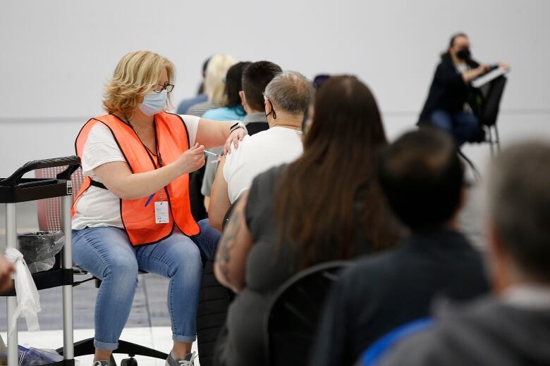 A person in an orange vest administers a needle to a person sitting in a room full of other people.