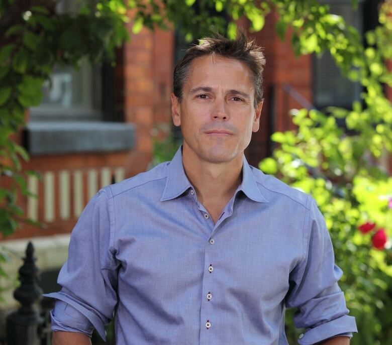 A person stands in front of trees and a house.