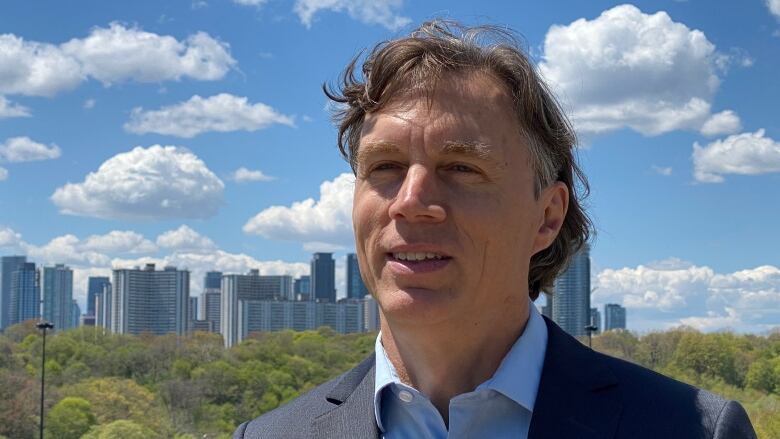 A man in a suit jacket and shirt stands in front of a skyline partially shrouded by trees