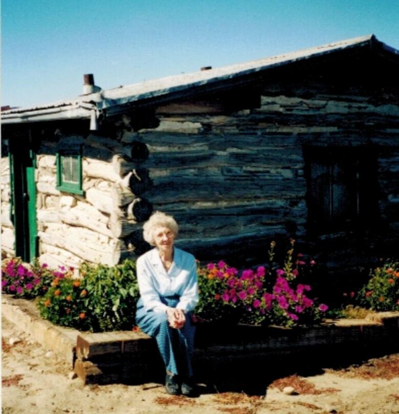 An elderly woman sits outside of a log home.