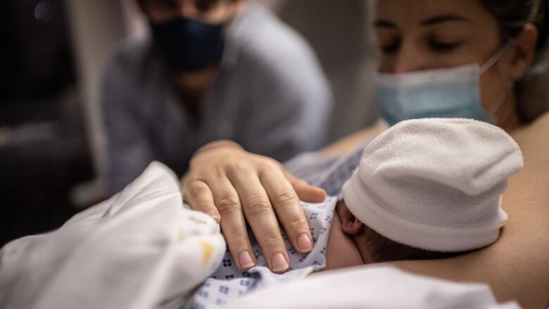 A newborn baby snuggles into their mother in a hospital bed. Their father looks on in the background. 