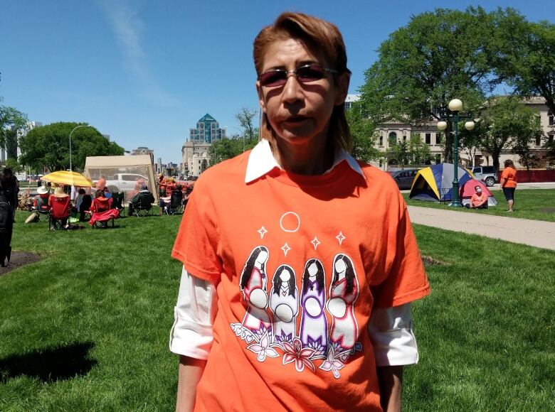 A woman with short red hair is pictured in an orange shirt that has an illustration of four women on it. Behind her are a number of tents and people.