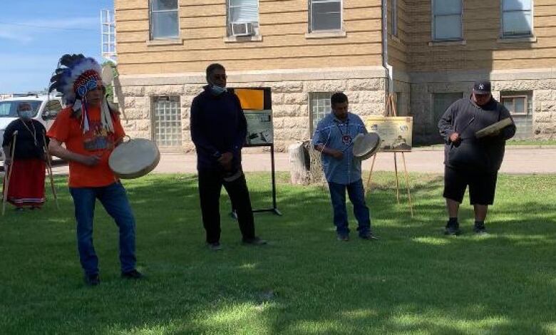 Four people in front of a brick building, including three who hold drums, with others standing in the background.