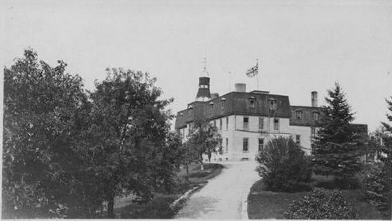 Old picture of a building up a road flanked by trees.