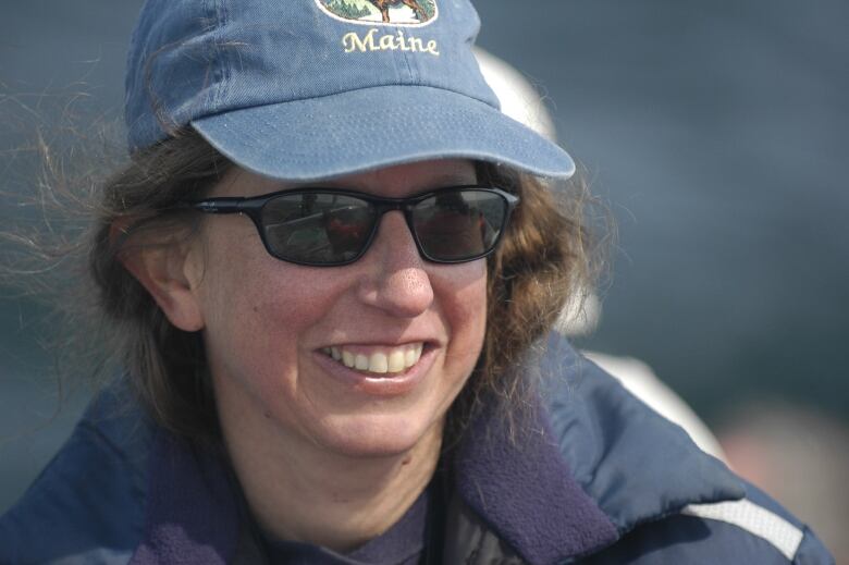 A woman in aball cap and sunglasses is outside and smiling.