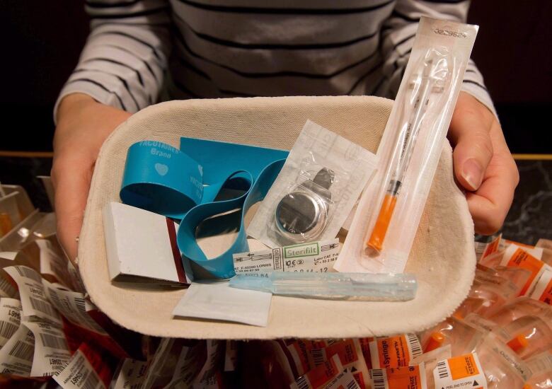 Two hands hold a tray containing sterile needles, drug test strips, rubber bands, alcohol swabs and a book of matches. 