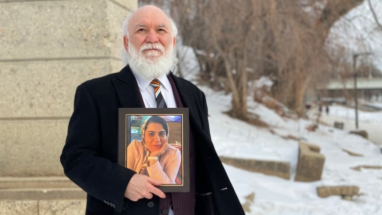 A man with a white beard and wearing a long black coat stands outside in winter, holding a photo frame.