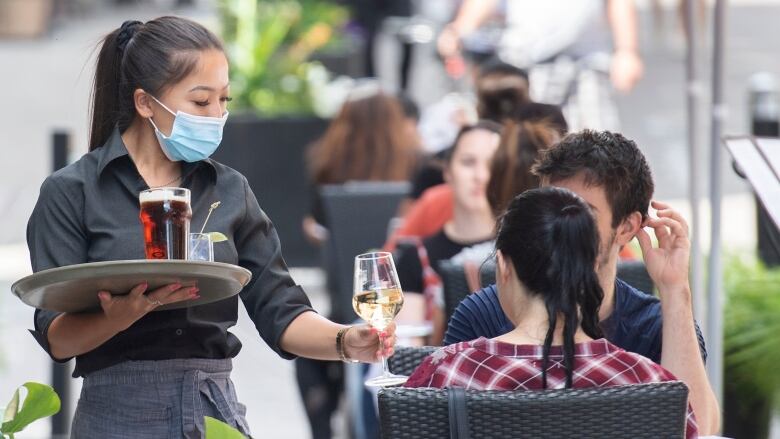 A sever wears a blue facemask and black uniform. She is serving a table of two people a glass of white wine and a beer. 