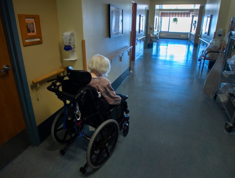 A woman with white hair sits in a wheelchair in the hallway of a seniors' residence.