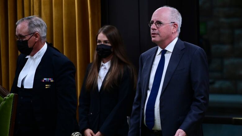 A man in a black suit and tie stands in the aisle of the House of Commons.