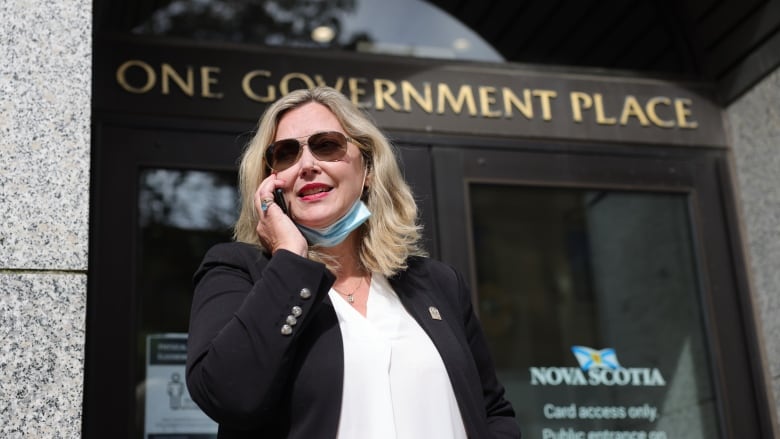 A woman with blonde hair wears a black blazer with a white shirt and sunglasses. She is standing outside in front of a building.