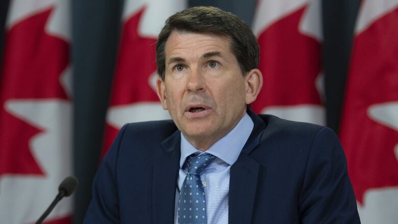 A man in a navy suit sits in front of a Canadian flag.