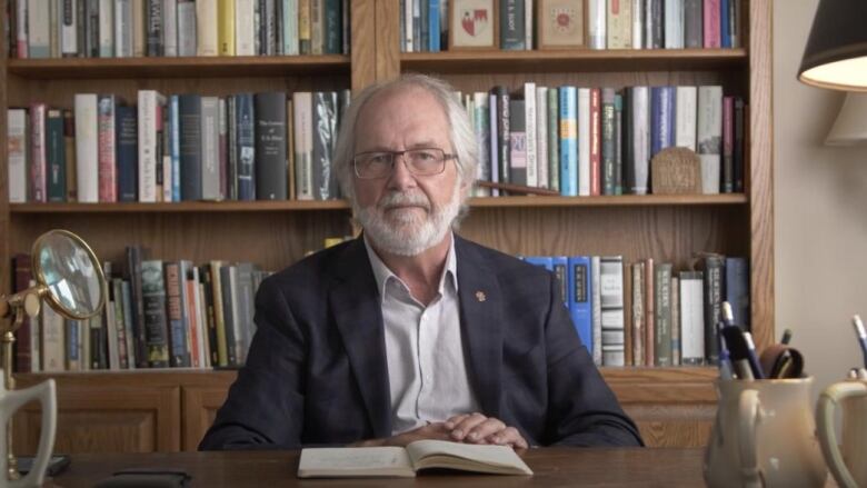 A man with white hair wearing glasses sits at a desk facing the camera. He's wearing a blue suit and is sitting in front of a bookshelf with lots of books.