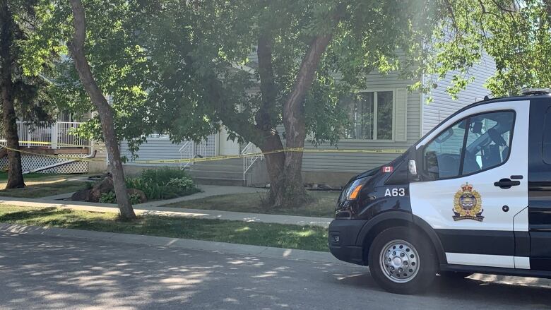 A marked police van parked on a leafy street, with yellow police tape stretched between boulevard trees.