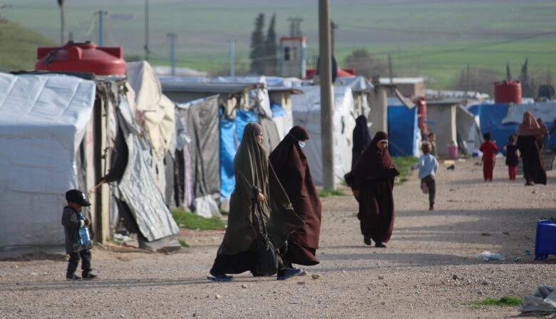 three women in head to toe Islamic coverings walk among children on a gravel road flanked by tents