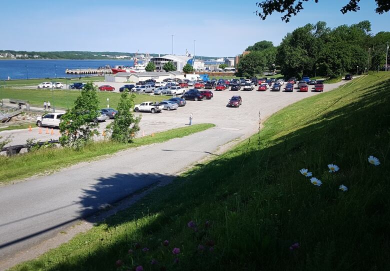 A gravel parking is shown on land surrounded by green grass next to a waterfront harbour.