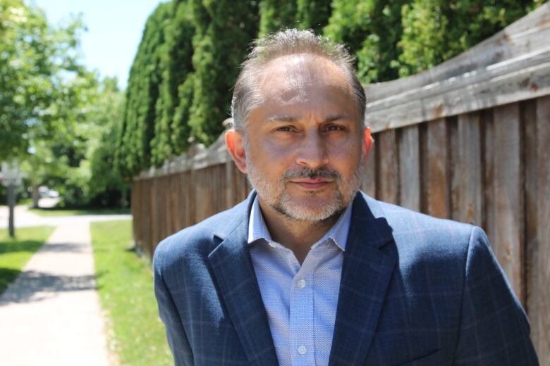 A man with grey hair and a beard in a light blue shirt and navy blazer stands on a sidewalk with a long wooden fence strethcing behind him. 