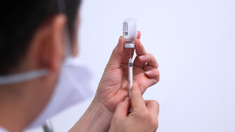 A person wearing a medical mask draws liquid from a vial with a syringe. Their face is visible from profile.