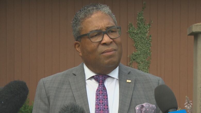 A Black man is seen wearing glasses and a grey suit. He is standing in front of several mics.