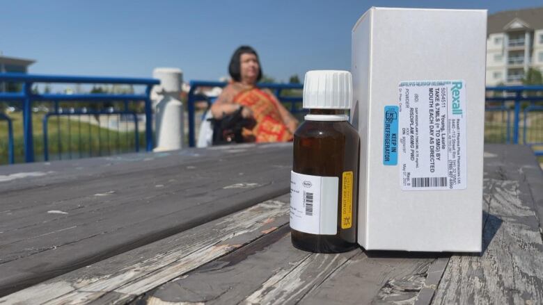 A woman sits behind a picnic table with a vial and box in the foreground.