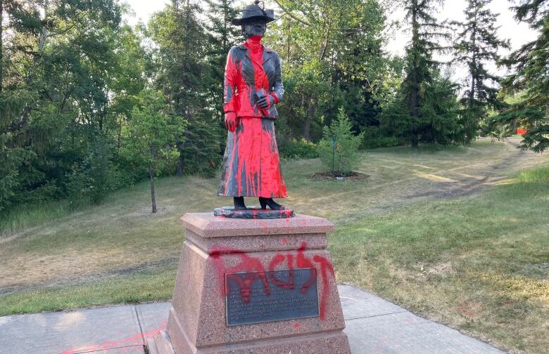 A statue of a woman in a park is defaced with red paint.