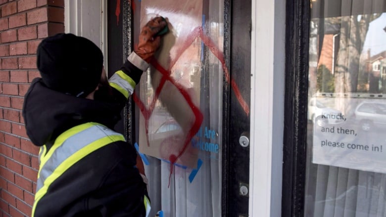 A graffiti removal worker cleans anti-Semitic graffiti, including a swastika, that was spray painted on the door of The Glebe Minyan in Ottawa, on Tuesday, Nov. 15, 2016. The federal government is making it easier for religious and cultural groups at risk of hate crimes to improve security in and around their buildings.