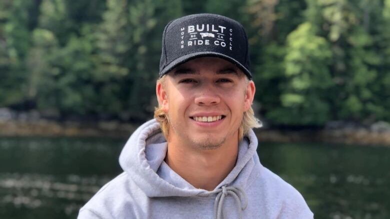 A photo of a smiling young man in a black ball cap and grey hoodie.