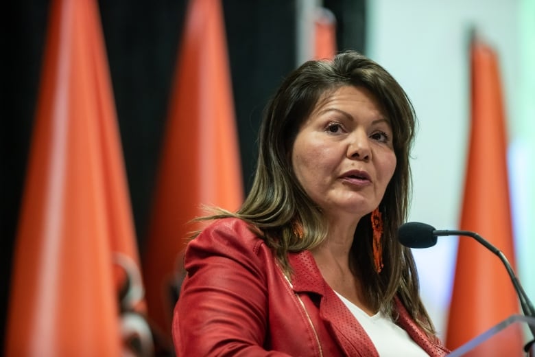 A woman in a red blazer sits at a table with orange flags behind her.