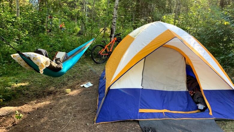 A blue tent with yellow trim sits in a wooded campsite. To the left of it, tied between two trees, is a teal hammock with a person inside reading a book. Their face is not visible.