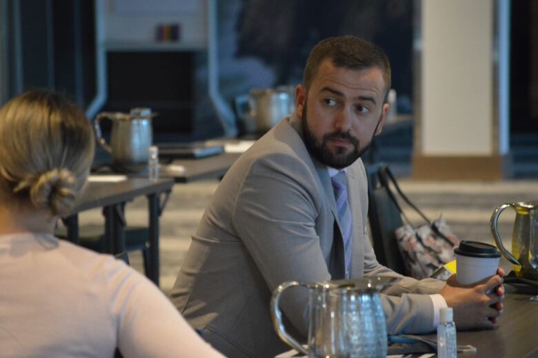 A bearded man in a suit leans forward on a table, his handles clutching a paper coffee cup. 