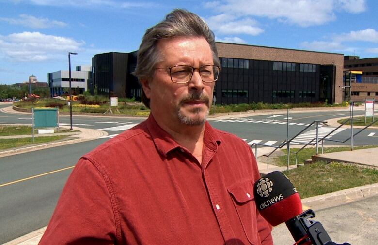 A man is standing outside a large building. He has glasses and the wind is blowing his hair. 