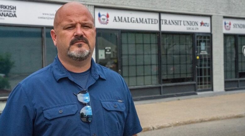 A man with a goatee in a blue shirt stands outside in front of a building with a sign reading, Amalgamated Transit Union