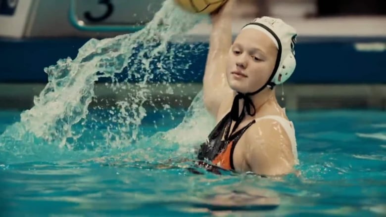 A woman in a white swim cap holds a ball behind her head while in a pool.