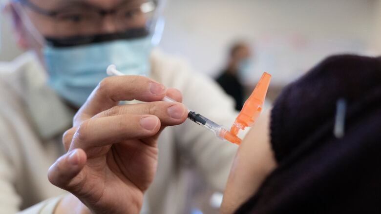 A closeup of a male medical professional in a white coat and blue medical mask injecting a COVID-19 vaccine in a person's upper left arm.