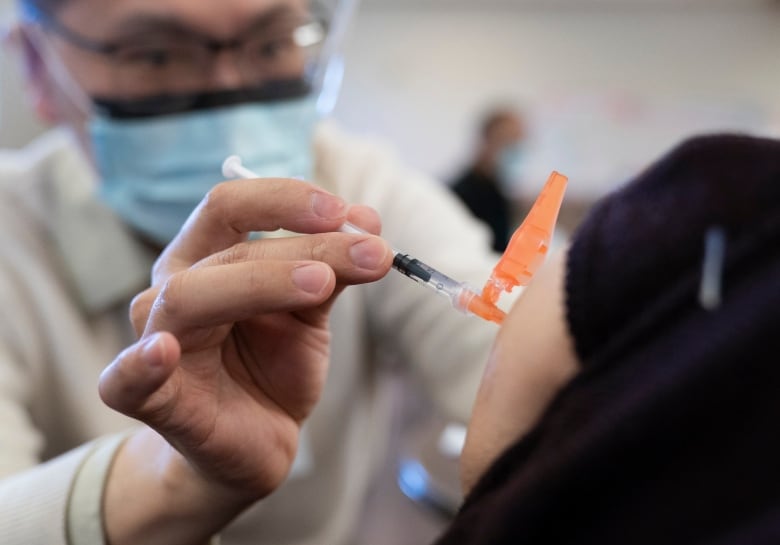 A closeup of a male medical professional in a white coat and blue medical mask injecting a COVID-19 vaccine in a person's upper left arm.