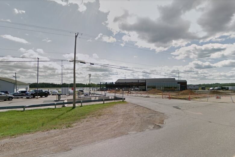 Wide pavement roads and electrical wires hang over a long, short building in the distance. The sky is blue with several grey clouds. 