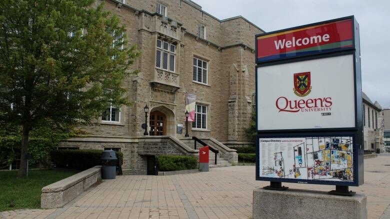 A sign says 'Welcome - Queen's University' in front of a stone building.