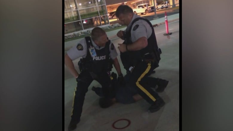 Two uniformed RCMP officers kneel on a man lying on a sidewalk, with one of the officers' knees on the man's neck.