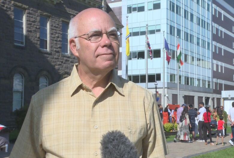 A man with glasses and a yellow shirt listens to a reporter's question near an outdoor rally. 