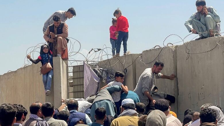 Men, women and children clamber over a concrete wall topped with razor wire. 