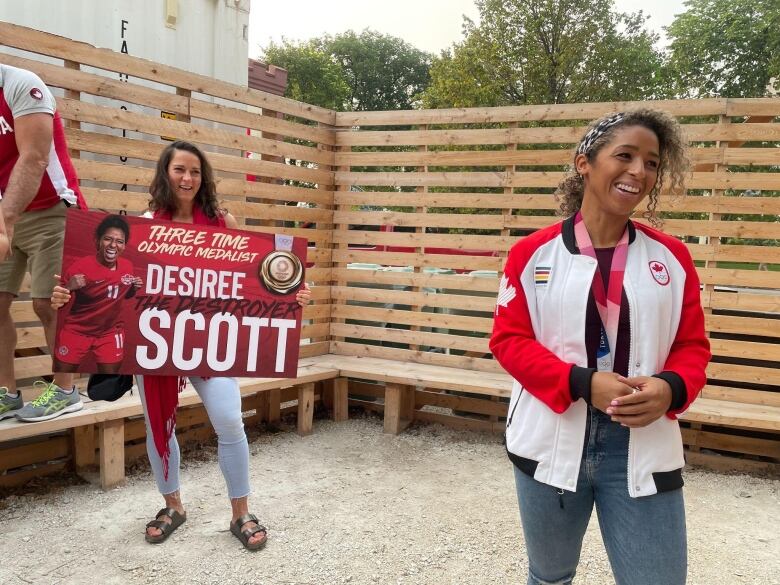 A smiling woman stands outside next to another person holding a sign reading 