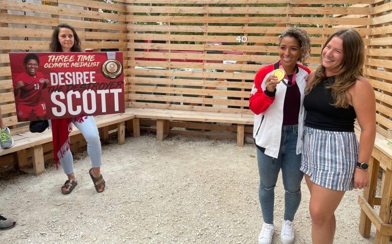 A woman smiles as she holds up a gold medal, posing next to a smiling young woman.