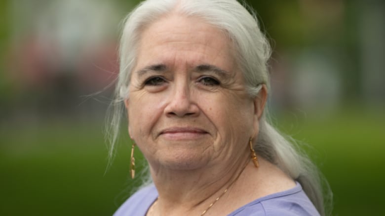 A woman with grey hair and gold earrings is smiling. She is wearing a purple shirt. 