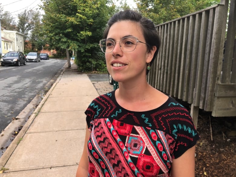 A woman wearing a bright patterned shirt and glasses stands on the sidewalk by a Halifax street.