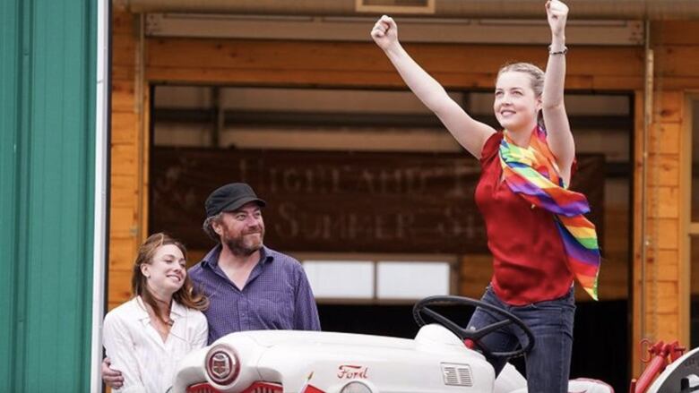 A young girl wearing a rainbow scarf stands on a tractor, raising her arms in the air triumphantly. Two adults stand nearby, looking at her lovingly.