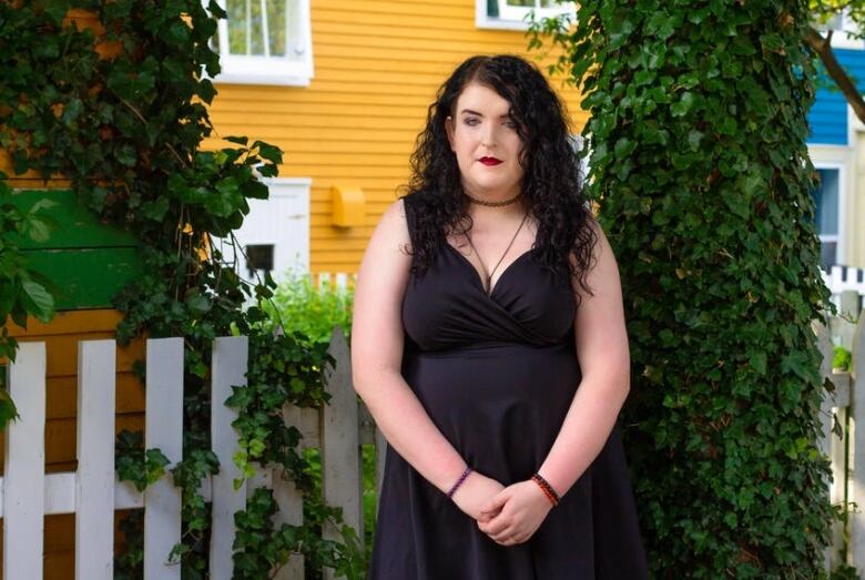 A person with dark hair in a dark dress stands next to a shrub.