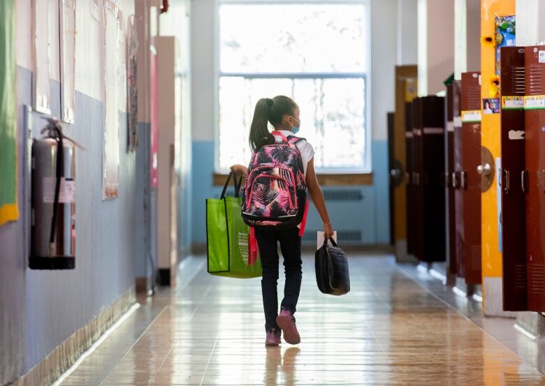 A young girl walks down the empty hallway of a school, past rows of lockers, carrying a large backpack, lunch box and recyclable shopping bag.