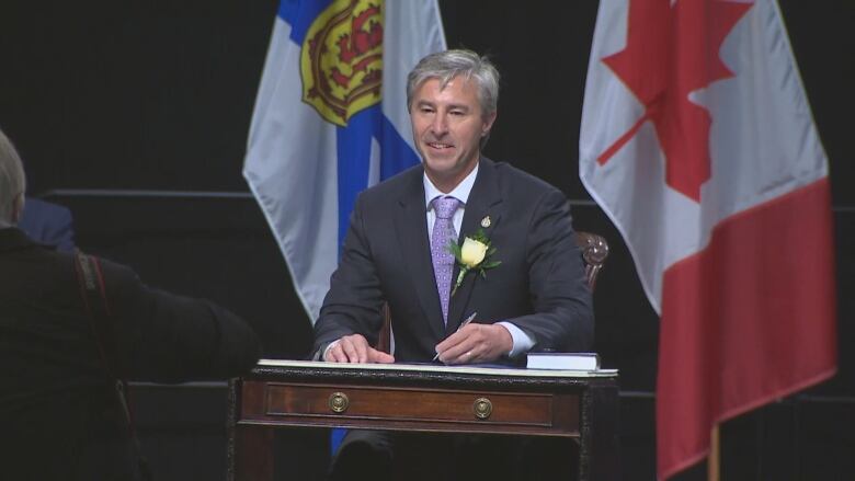 A white man with grey hair is seen sitting at a desk, pen in hand, wearing a dark grey suit