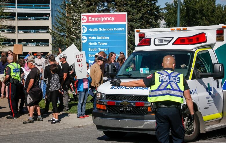 Police direct an ambulance past a crowd of people, one of whom is carrying a sign that says Just say no.