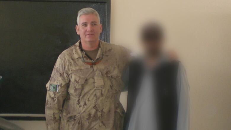 A military officer in uniform stands with his arm around a man whose face is blurred to protect his identity.